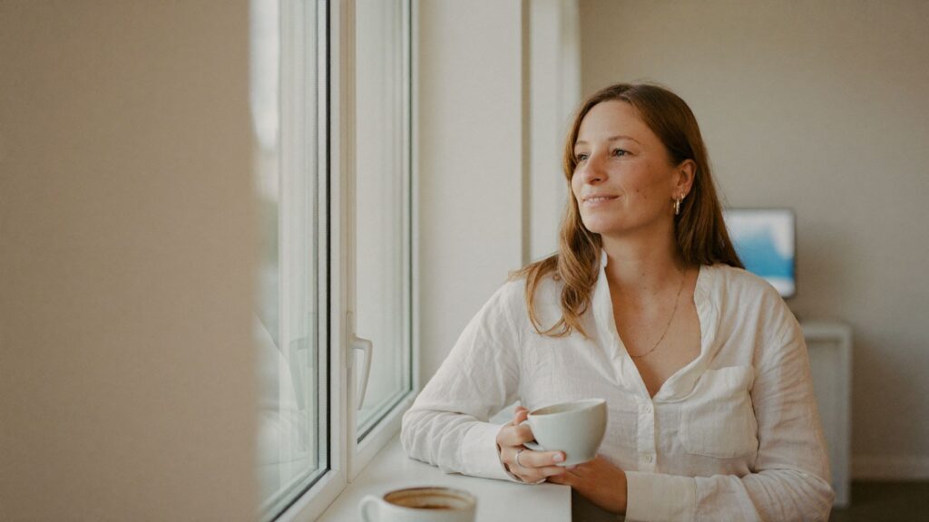 Eine Praxisinhaberin steht mit einem Kaffee am Fenster ihrer Praxis und blickt hinaus.